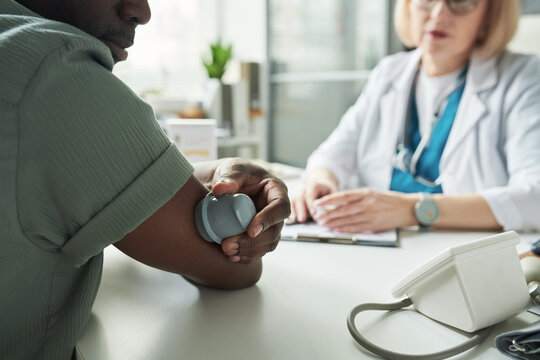 Black middle aged man checking blood glucose with medical device while sitting across from Caucasian middle aged female doctor in medical office during diabetes consultation