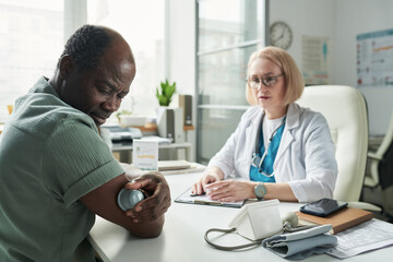 Obraz premium Middle aged Black man checking blood glucose with glucometer while sitting across from middle aged Caucasian female doctor in medical office during diabetes consultation