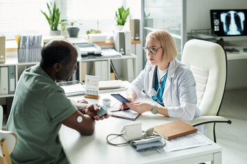 Fototapeta premium Middle aged Black man sitting across from Caucasian female doctor discussing diabetes management during medical consultation in modern office, blood glucose monitor and documents on desk