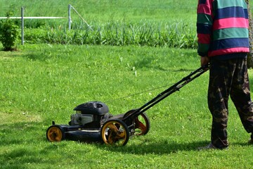 Fototapeta premium Person mowing green lawn with an old gasoline push mower. Only lower body visible. Summer outdoor garden work, casual clothes and camo pants. Concept of yard maintenance and countryside living.