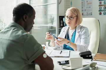 Middle aged Caucasian female doctor explaining insulin pen to middle aged Black man with glucose...
