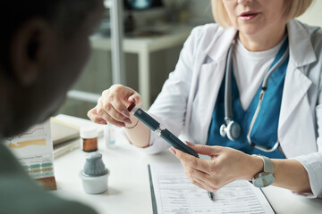 Caucasian middle aged female doctor explaining use of blood glucose meter to Black middle aged man during diabetes consultation in medical office, hands holding device over patient chart