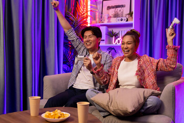 Couple gamer raising fist up holding joysticks be winner together complete level sitting sofa playing video game with front snack and drinks at neon light color living room at modern home. Infobahn.