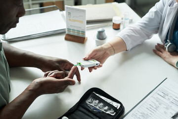Middle aged Black man having blood glucose level checked by female doctor in medical office, doctor using glucometer on patient finger, diabetes management consultation