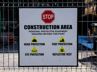 Safety sign at a construction site, listing required personal protective equipment including helmet, boots, glasses, and reflective vest. Warning for all entering the restricted area.