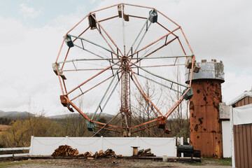 Rusted abandoned Ferris wheel frame behind white fence with stacked firewood in rural overcast...