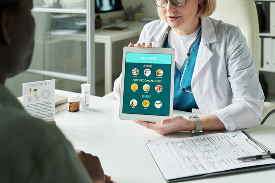 Middle aged Caucasian female doctor showing digital tablet with diabetes dietary recommendations to middle aged Black man during medical consultation in modern clinic setting