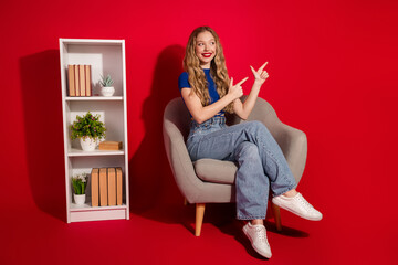 Charming blonde girl in casual outfit pointing with hands inside modern room with bookshelf against bright red background