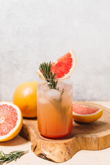 Grapefruit cocktail with rosemary garnish in a glass, served on a wooden cutting board. Vibrant,...