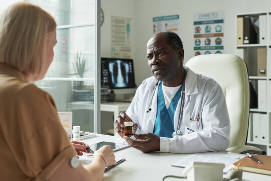 Senior Caucasian woman with glucose monitor on arm consulting Black male doctor in medical office, doctor holding prescription bottle and discussing diabetes treatment options - Powered by Adobe