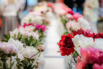 Rows of vibrant peony flowers in various shades of pink, red, and white are displayed in vases on tables at an indoor floral exhibition.