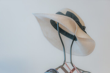 A woven straw hat hangs on a black coat rack with light peach accessories, including a scarf and beads, against a softly lit pale background.