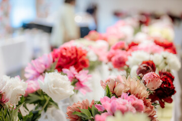 Rows of vibrant peony flowers in various shades of pink, red, and white are displayed in vases on tables at an indoor floral exhibition.