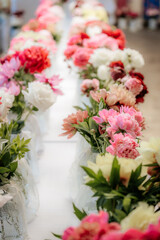 Rows of vibrant peony flowers in various shades of pink, red, and white are displayed in vases on tables at an indoor floral exhibition.