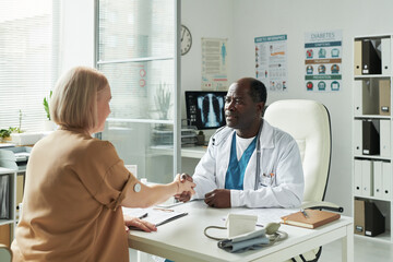 Obraz premium Middle aged Caucasian woman with glucose monitor on arm visiting Black male doctor in medical office, shaking hands across desk with medical documents and diabetes chart visible