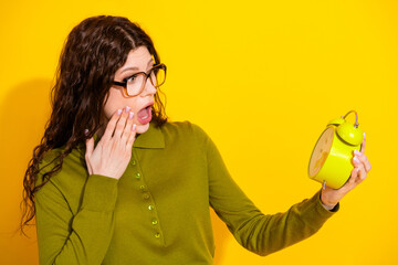 Surprised young woman in green sweater with alarm clock isolated on vibrant yellow background