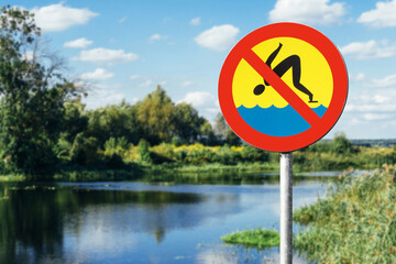 No jumping into water sign. Warning symbol near lake. Shallow water danger. Risk of injury from diving. Red and yellow circle sign. Nature background with river and trees. Safety alert outdoors.