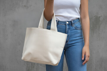 Short canvas tote bag mockup held by woman in jeans and white top in natural light setting with concrete wall background for realistic promotional design