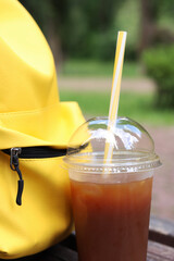 Coffee with juice in a disposable plastic cup with a straw. Coffee with orange juice, syrup and ice. Refreshing drink near a yellow backpack, rest for walkers in hot weather. Takeaway drink. Bumble