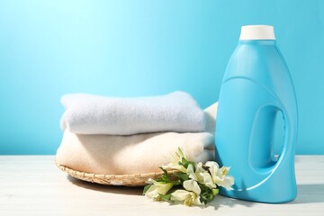 Bottle of fabric softener, clean clothes and lily flowers on white wooden table against light blue background