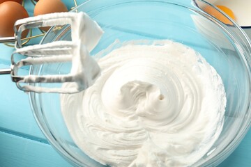 Bowl with whipped cream, hand mixer and ingredients on light blue wooden table, closeup