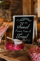 Rustic candy buffet with “Love Is Sweet Please Take A Treat” chalkboard sign, pink candies and rock sugar sticks in glass bowls on a burlap-covered table