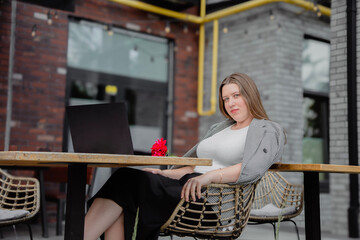 Young woman in gray jacket sitting at a table in a street cafe with a laptop and a red rose, looking at the camera. Business atmosphere, relaxation.