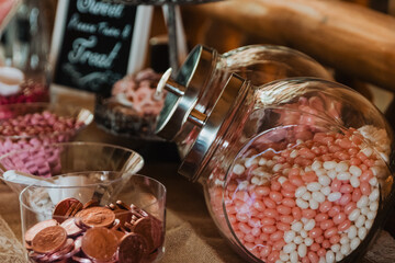 Tilted glass jar of pink and white jellybeans beside bowls of copper chocolate coins and pink candies on a burlap-covered candy buffet with “Love Is Sweet” chalkboard sign