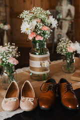 Pair of pink glitter ballet flats and brown leather oxfords displayed on a burlap-covered table beside mason jars of daisies, carnations and baby’s breath in a rustic interior