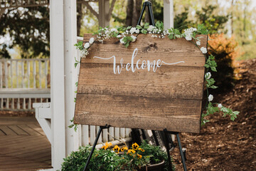 Rustic wooden welcome sign on black easel adorned with white rose blooms, baby’s breath and...
