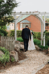 Bride in lace gown holding a bouquet and groom in dark suit walk together under a white garden arbor along a gravel path toward a rustic lodge