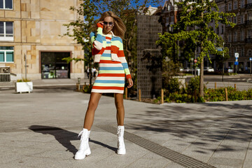 Pretty woman posing in a colorful striped dress and white boots on a sunny city street, showcasing...