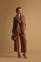 A fashionable woman wearing a brown linen pantsuit with relaxed tailoring and structured lapels, posing confidently against a neutral background. Ideal for editorial and commercial use.