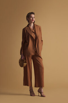 A fashionable woman wearing a brown linen pantsuit with relaxed tailoring and structured lapels, posing confidently against a neutral background. Ideal for editorial and commercial use.