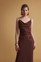 Confident fashion model in a brown satin slip dress and golden belt, posing in studio for a...