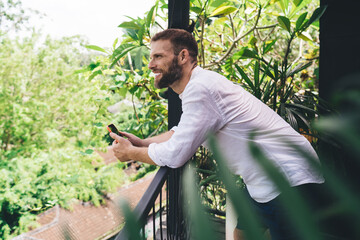 Freelancer leans outdoors holding smartphone, smiling while engaged online. Illustrates human...