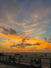 Sunset at the beach, orange and blue sky with some clouds. A few people are gazing on the sand. At...