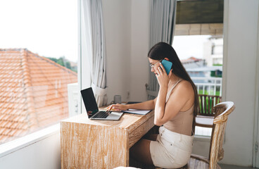 Freelancer woman multitasks between smartphone call and laptop, showing digital communication, tech efficiency and remote career multitasking in bright home office.