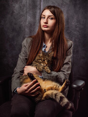 Obraz premium Classic beauty portrait of a young girl in grey business suit holding cat on grey painted background. Fashion and beauty. Model has long hair and calm confident face. Feminine soft pose. Relaxed mood