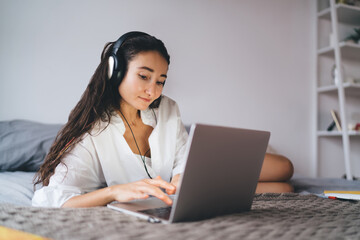 Focused woman types on laptop while listening through headphones, immersed in online learning or...