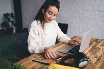 Young adult female freelancer checking smartphone next to laptop, multitasking efficiently with...
