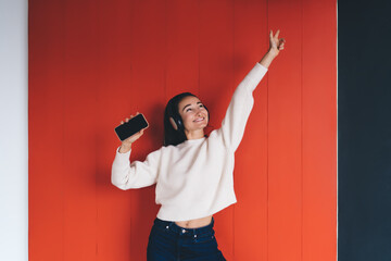 Charming woman dancing in headphones, smiling, holding phone, expressing lightness and musical...