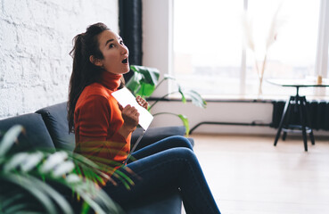 Surprised young woman holding laptop while sitting on sofa, expressing emotional reaction to...