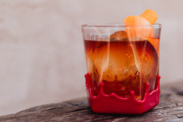 Close-up of red-wax-dipped rock glass holding amber cocktail with spherical ice and curled orange-peel garnish on weathered wooden table