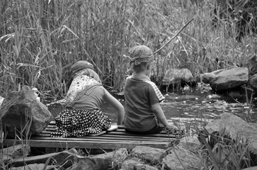 Horizontal black and white photo of two children sitting on a wooden bridge near a small stream,...
