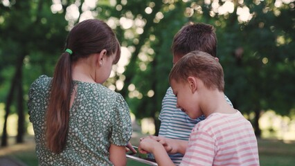 Children friends, school students play on tablet on street in park. Children friendship. Modern gadget kid. Happy child boy, girl play with smart tablet. Children communicate with each other on street