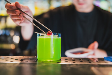 Bartender placing red garnish into a green cocktail with tongs on a bar counter. Close-up shot with blurred background, showcasing drink preparation.