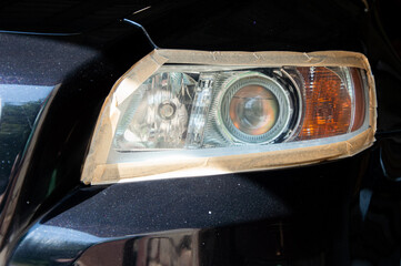 A person is carefully polishing a car headlight, using masking tape to protect the surrounding area during the automotive maintenance process