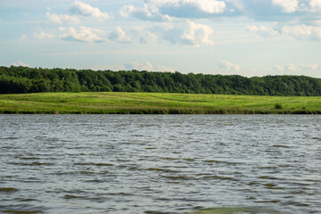 A vibrant green field borders a calm river, with dense trees lining the shore under a partly cloudy...