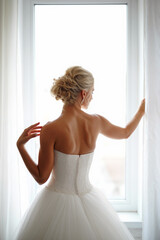 A bride in a strapless wedding gown stands by the window, softly touching the curtains while looking outside. The golden light enhances the serene moment. Generative AI.